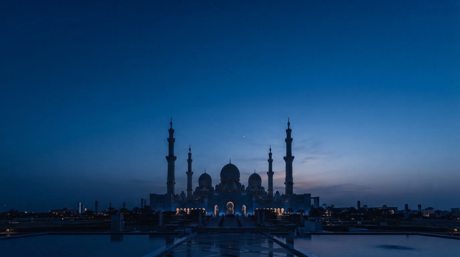 A mosque with four minarets, lit from within, under a deep blue sky before dawn.
